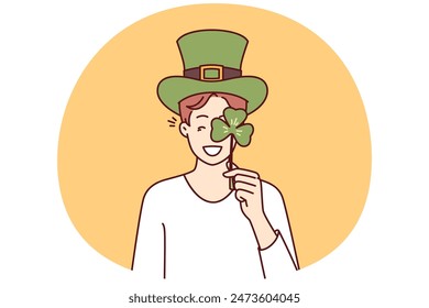 Happy man with St Patrick Day celebration accessories wearing green hat and covering eye with clover petal. Positive guy invites to traditional festival on eve of Irish holiday St Patrick Day