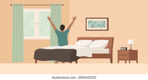 Happy man waking up in the morning, he is sitting on the bed and stretching his arms