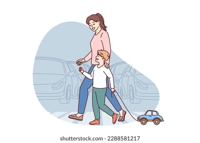 Happy mom and child going along pedestrian crossing across road walking through summer city. Little boy with toy car walks with nanny teaching how to cross intersection road correctly 