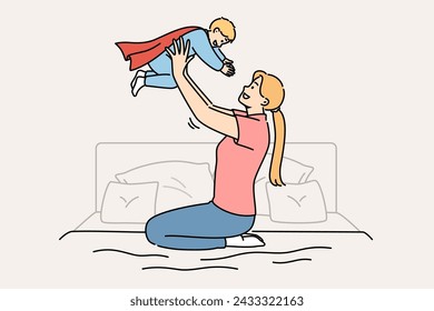 Happy mother lifts up newborn son in superhero costume, sitting on bed in bedroom. Caring casual mother plays with child, motivating son to achieve ambitious goals and success in life