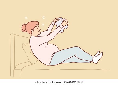 Happy pregnant woman sits on bed holding small t-shirt for baby in hands and rejoices at imminent arrival of son. Pregnant girl who will soon become mother and prepares clothes for newborn in advance