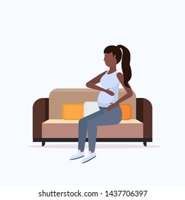 happy pregnant woman sitting on couch african american girl holding her bump girl pregnancy matherhood concept full length flat
