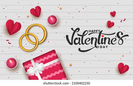 Happy Valentine's Day Font With Top View Of Golden Couple Rings, Gift Box, Paper Cut Hearts And Lit Tea Candles On White Wooden Texture Background.