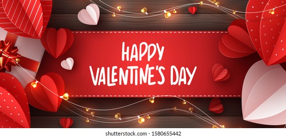 Happy Valentines day holiday banner. Decorative paper cut hearts, gift box, garland with handwritten lettering text on brown wooden background. Vector illustration.