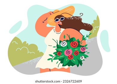 Happy woman with bouquet of wild flowers walks along sidewalk in summer park and holds straw hat with hand. Positive girl with flowers given by boyfriend on date, enjoying sunny weather