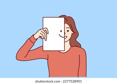 Happy woman covers half of face with paper with smiling emoticon, wanting to share good mood and positive with people around. Kind girl carelessly looks at camera enjoying life and enjoying moment 