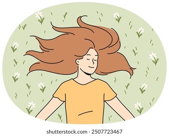 Happy woman with long hair lies on grass in park with eyes closed and smiles rejoicing in sunlight. Positive young woman in casual t-shirt enjoying good summer weather lying on lawn with flowers