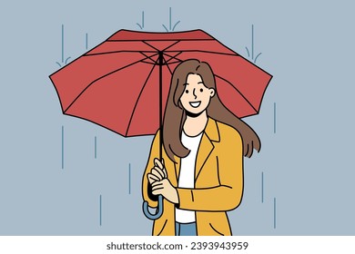 Happy woman with umbrella walks in rain along autumn street and looks at screen with wide smile. Cheerful girl dressed in casual clothes uses umbrella to avoid getting wet and catching cold.