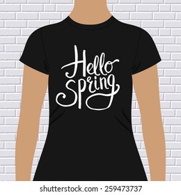 Hello Spring simple t-shirt design with flowing white script text on the chest of a black t-shirt modeled on a person against a white brick wall