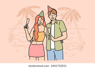 Pareja feliz de luna de miel bebiendo champán, de pie en la playa de resort soleado, con gorras de cumpleaños. Chico y chica románticos celebran el comienzo de la luna de miel en la orilla del océano con palmeras