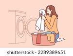 Housewife woman sits near washing machine and inhales fragrant smell freshly washed towel after using good laundry detergent. Girl takes out washed clothes from washing machine putting them in bowl 