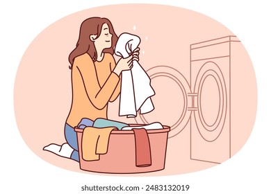 Housewife woman sits near washing machine and inhales fragrant smell freshly washed towel after using good laundry detergent. Girl takes out washed clothes from washing machine putting them in bowl