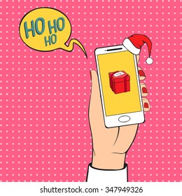 Human hand holding a white mobile phone with gift icon on the screen. Vector illustration. Christmas background in retro style. Santa red hat and speech bubble with text.
