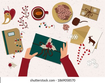 Las manos humanas empacan un regalo de Navidad. La vista desde arriba Mentiras planas. Una taza de té, galletas, postales, dulces y accesorios están repartidos por todas partes. Ilustración vectorial.