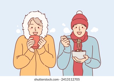 Hungry friends eat lunch standing under snow in winter clothes, trying to stay warm during december trip. Tourists drink coffee and eat noodles located outdoors, enjoying trip to winter resort