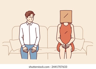 Indecisive man sits near woman with paper bag on head and is afraid of becoming acquainted. Indecisive guy sits on sofa with girlfriend, and is stressed due to timidity interfering with relationships