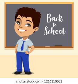 An Indian school boy in his uniform is standing in front of a blackboard in his classroom