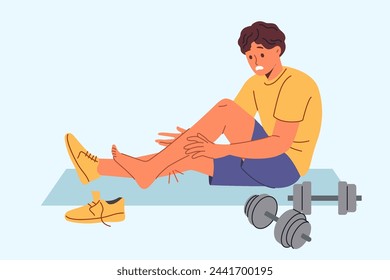 Injured sportsman sits on mat for sports and gymnastics near dumbbells, after stretching muscles on legs. Injured athlete experiences pain and agony after careless use of sports equipment