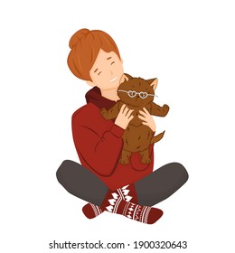 Isolated Red girl in hoodie and knitted socks holds a cute fluffy brown cat or kitten. She and pet are getting ready for Valentine's Day. The cat has white heart-shaped glasses on its face