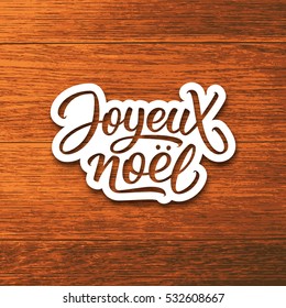 Joyeux Noel text on paper label with hand lettering over wood background. Merry Christmas sticker or greeting card vector design template with french inscription