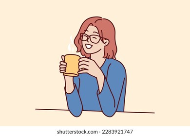 Joyful woman holding coffee mug sitting at table and smiling remembering happy moments from life during lunch break. Girl in casual clothes drinks hot tea or coffee to gain vigor and energy
