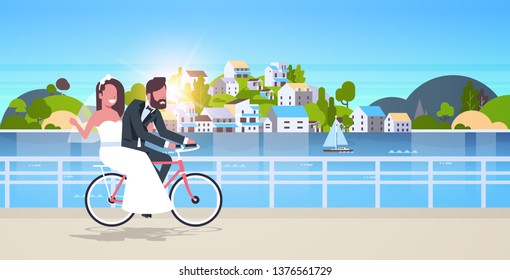just married man woman riding bicycle romantic couple bride and groom cycling bike having fun wedding day concept mountain city island sunset background full length horizontal flat
