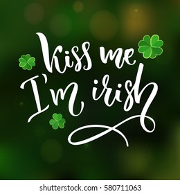 Kiss me, I'm irish. Fun St. Patrick's day quote for t-shirts and cards. Brush lettering design with clovers at green blurred background.