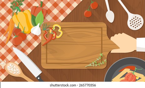 Kitchen wooden worktop with cook's hand holding a chopping board, kitchen tools and vegetables all around, top view