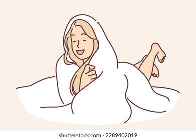 Laughing woman wrapped in blanket lies on bed not wanting to get up and go to work or college. Positive blonde girl spends evening lying under warm blanket and enjoying comfort cozy atmosphere. 