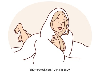 Laughing woman wrapped in blanket lies on bed not wanting to get up and go to work or college. Positive blonde girl spends evening lying under warm blanket and enjoying comfort cozy atmosphere.