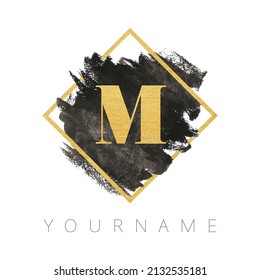 Letter M logo design. Black watercolor brush stroke with gold letter and gold square frame.