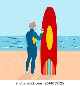 A lifeguard in a blue wetsuit stands next to a red and yellow rescue board on a sandy beach with the ocean in the background.