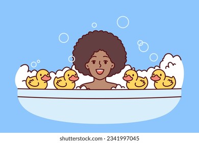 Little African American girl bathes in bubble bath with rubber ducks and looks at screen smiling. Cheerful child, surrounded by soap bubbles, bathes and enjoys performing useful hygiene procedures.