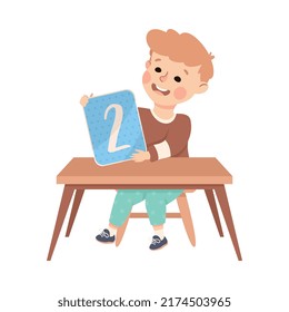 Little Boy with Number Two Sitting at Desk Showing Card with Numeral Vector Illustration