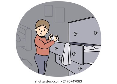 Little boy is standing near closet with clothes, looking at textiles with curiosity and studying world around. Childish pranks of funny boy throwing clothes from drawer making mess while playing