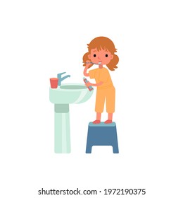 A little cute girl is brushing her teeth with a toothbrush at the sink, she is standing on a stool. The girl is holding a toothpaste in her hand. A glass of water on the sink. Morning hygiene. 