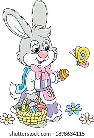Little Easter Bunny in a colorful holiday dress holding a small wicker basket of painted eggs and playing with a playful butterfly flittering around, vector cartoon illustration isolated on white