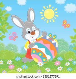 Little Easter Bunny and a colorfully decorated big egg among flowers and flittering butterflies on a green lawn on a sunny spring day, vector illustration in a cartoon style