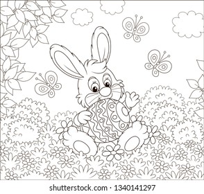 Little Easter Bunny sitting with a colorfully decorated big egg among flowers and flittering butterflies on a lawn on a sunny spring day, black and white vector illustration in a cartoon style