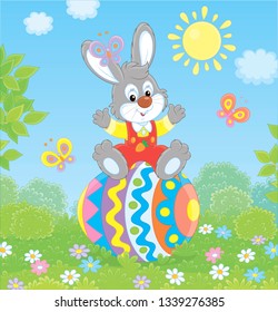 Little Easter Bunny sitting on a colorfully decorated big egg among flowers and flittering butterflies on a green lawn on a sunny spring day, vector illustration in a cartoon style