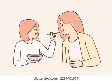 Little girl feeds mother with spoon during breakfast sitting at kitchen table spending time together. Mother and daughter having breakfast for concept of parenting and happy childhood or motherhood 