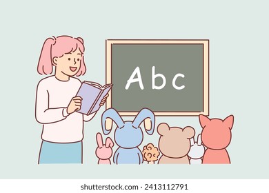 Little girl plays school teacher and reads book to toys sitting near blackboard with alphabet. Funny schoolgirl dreams of becoming teacher when she grows up to teach pre-teen children