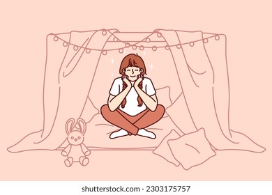 Little girl is sitting in home hut playing children games and dreaming of becoming princess with own castle. School-age girl smiles enjoying exciting game and cozy hut created from sheets and garlands