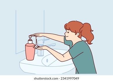 Little girl uses liquid soap to wash hands standing near sink and mirror in bathroom. Child follows hygiene rules with smile and washes hands with soap after coming home from school.