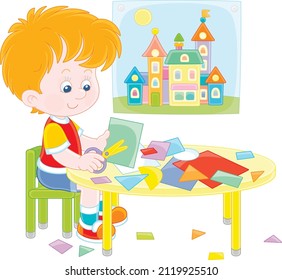 Little preschool child cutting outlines and figures from color paper with scissors and making a funny picture of a pretty toy town, vector cartoon illustration isolated on a white background