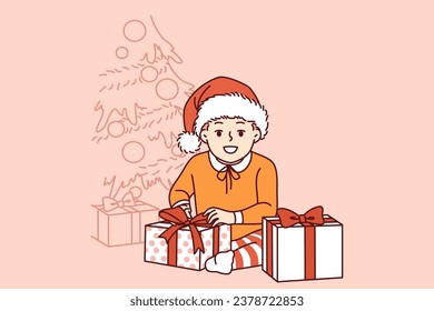 Little pre-teen boy sits near christmas tree and gifts from santa claus and smiling looks at screen. Kid rejoices at arrival of christmas or new year and large number of giftboxes from parents