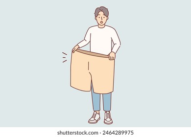 Abgefallener Mann mit großen Shorts in den Händen ist überrascht über die Größe der eigenen Kleidung aus der Vergangenheit. Ein Typ mit abgenommenem Gewicht rühmt sich der hervorragenden Ergebnisse von Diät und regelmäßiger Bewegung, die helfen, das Ziel zu erreichen