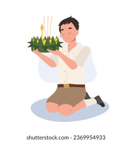 Loy Krathong Traditional Festival. Thai Student boy is sitting and hold krathong up to make a wish before float in river.