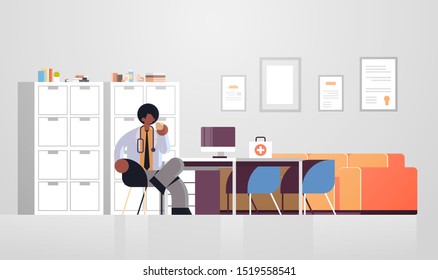 Male Doctor In White Coat Having Coffee Break Medicine Healthcare Concept African American Medical Worker Sitting At Workplace Modern Hospital Clinic Office Interior Full Length Flat Horizontal