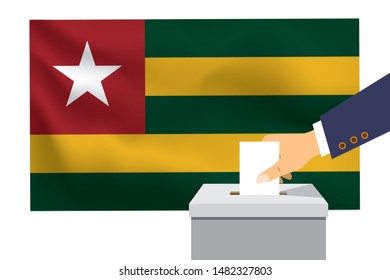 Male hand puts down a white sheet of paper with a mark as a symbol of a ballot paper against the background of the Togo flag. Togo the symbol of elections.